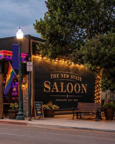 Concept rendering detail: the side-wall mural reading The New State Saloon, Premier Gay Bar, with a bench and a Y’all Means All chalkboard on the sidewalk.