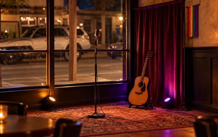 small performance corner by the front window, with a microphone stand, an acoustic guitar, a burgundy curtain and a Pride flag.